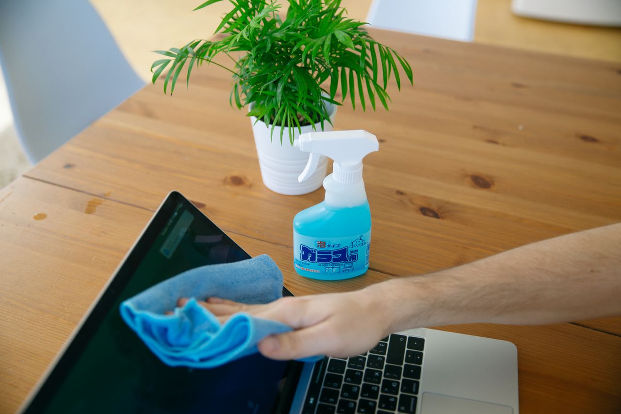 home-img Vue rapprochée d’une main nettoyant l’écran d’un ordinateur portable avec un chiffon bleu et un flacon pulvérisateur sur un bureau en bois.